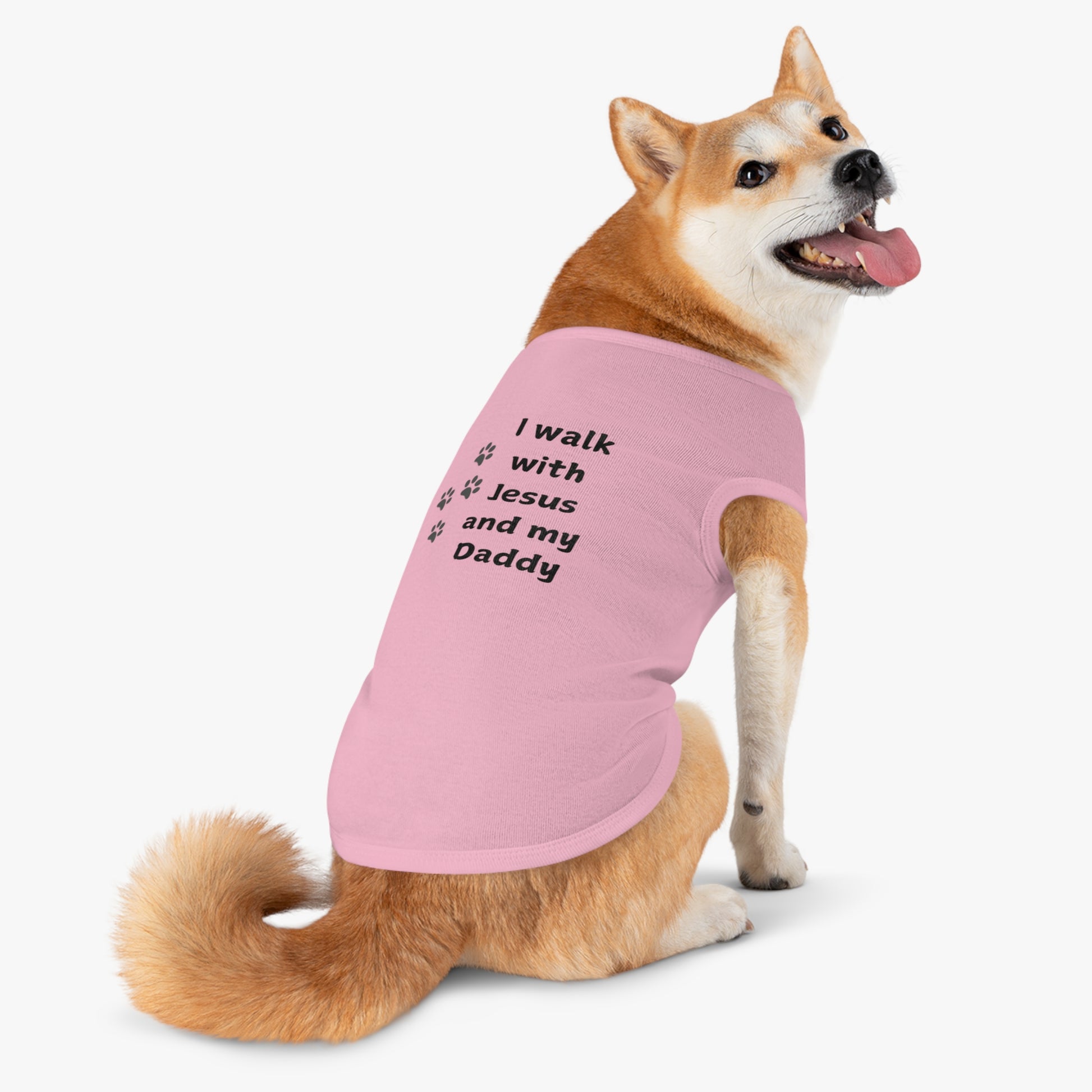 A brown and white dog wearing a pink t-shirt with the text "I walk with Jesus and my Daddy" printed in black, along with three paw prints.