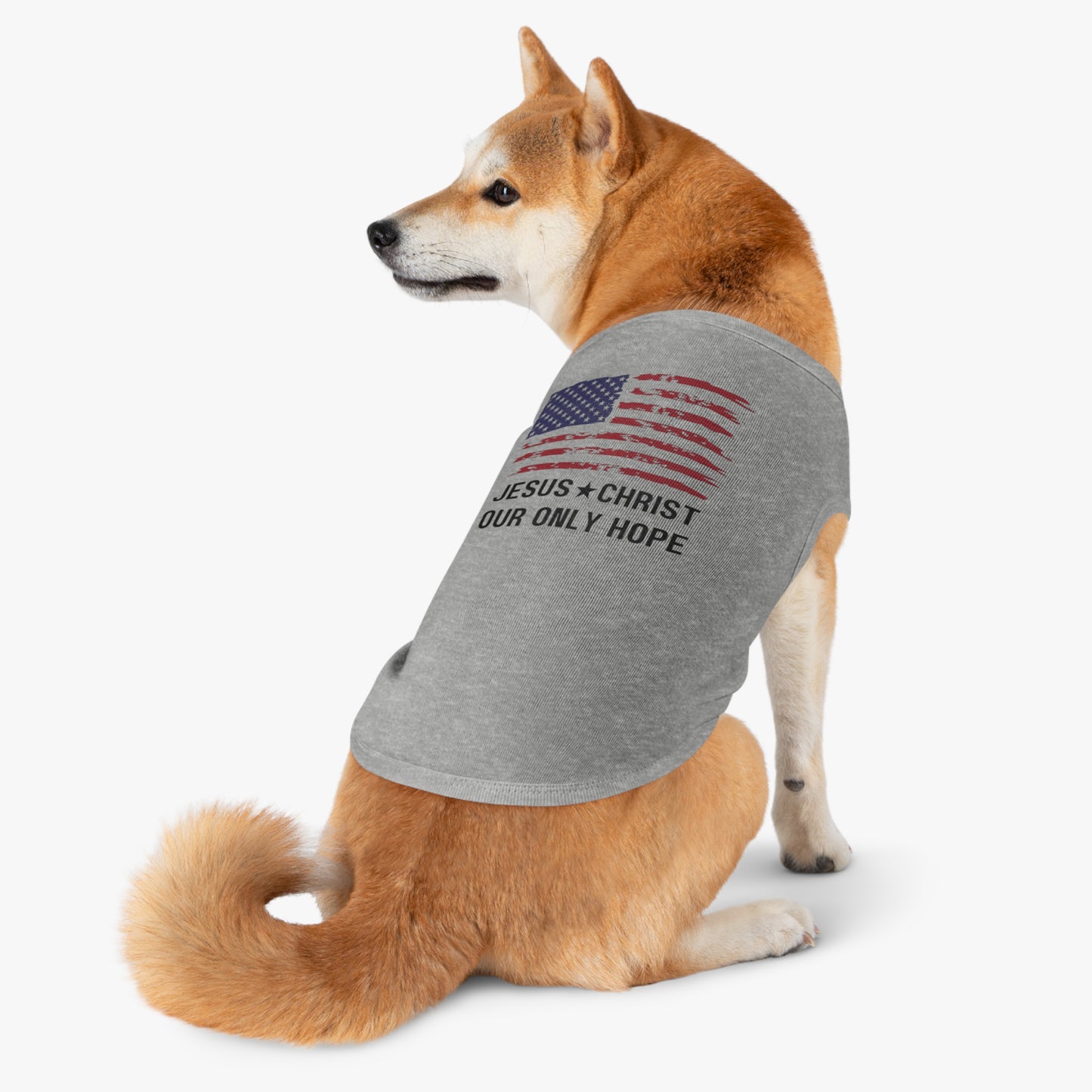 A small dog wearing a gray tank top with an American flag and the text "JESUS CHRIST OUR ONLY HOPE