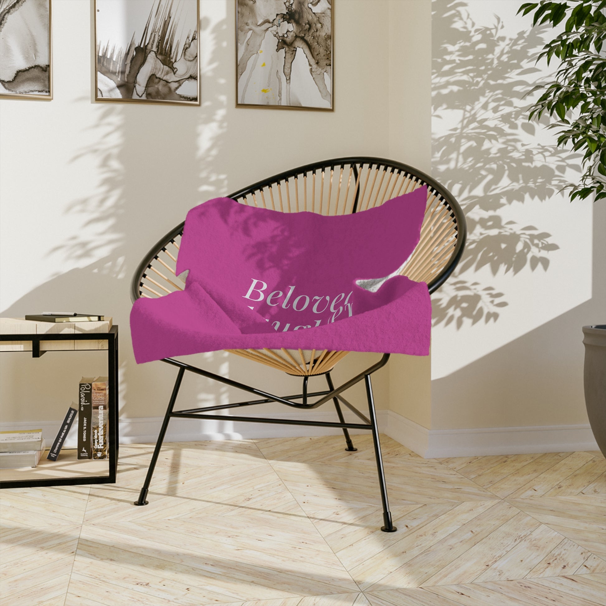 A pink throw blanket with the word "Beloved" printed in white lettering, resting on a stylish rattan chair with a black metal frame. The chair is positioned in a sunlit living room with artwork on the wall.